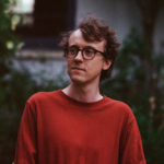 Pianist Joseph Havlat wearing glasses and a red jumper looking pensive.